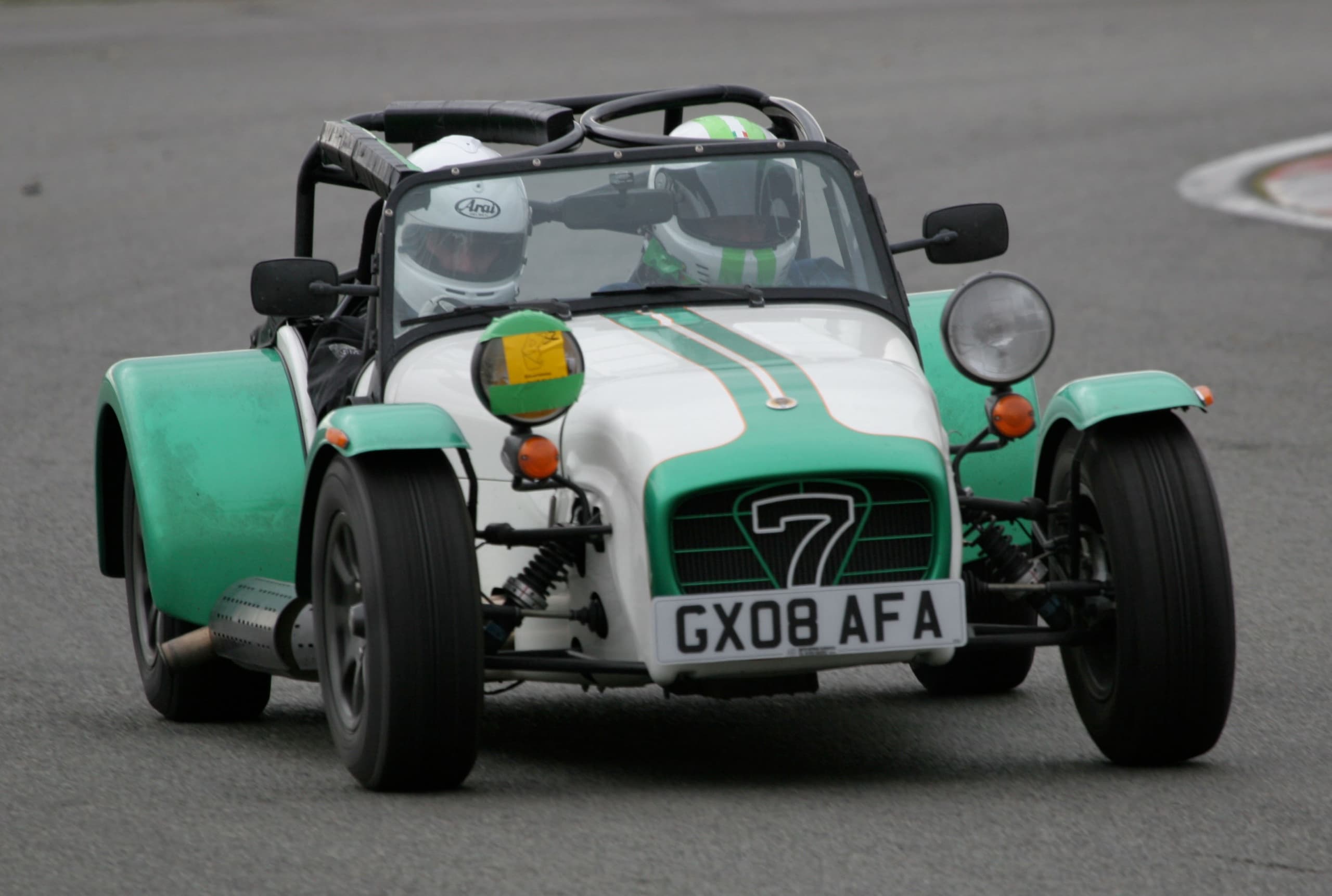 A green and white Caterham Seven with two helmeted occupants on a track day